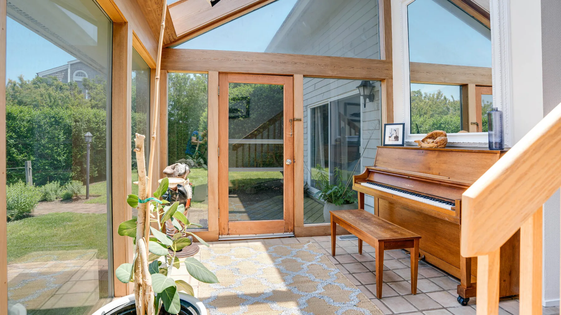 A bright sunroom with tall windows, a wooden piano, and plants.