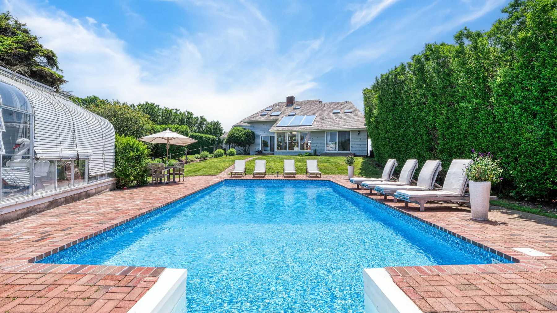 Una piscina al aire libre está junto a un patio de ladrillo, con una casa al fondo.