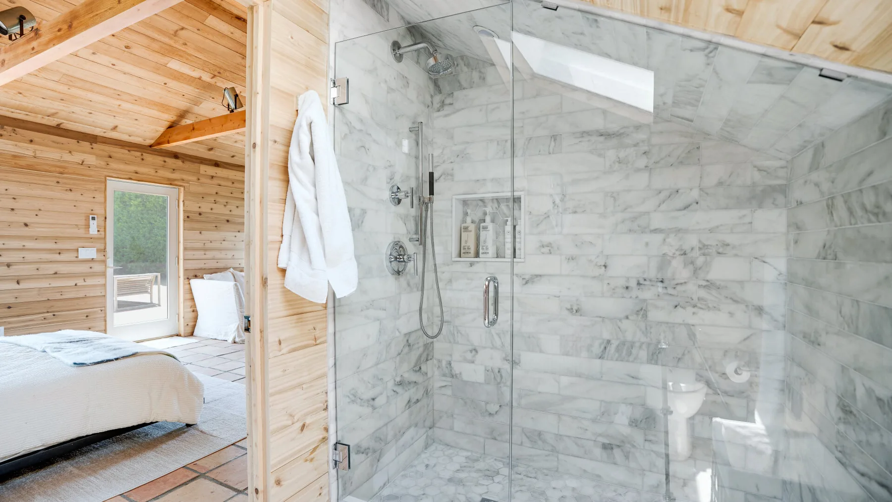 A bedroom beside a marble-tiled shower, with light wood walls and ceiling.