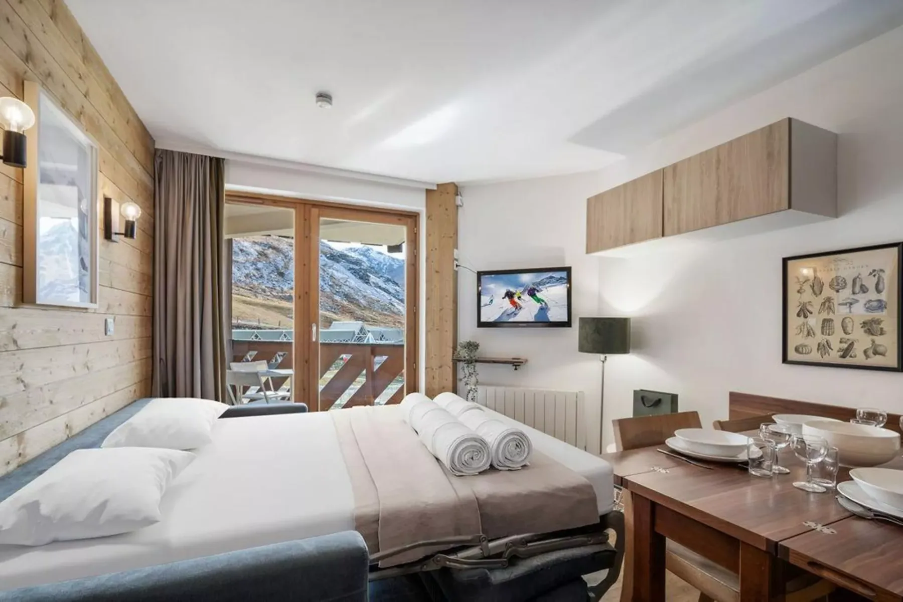 Dormitorio y comedor con una cama, una mesa y vistas a las montañas por las puertas de vidrio.