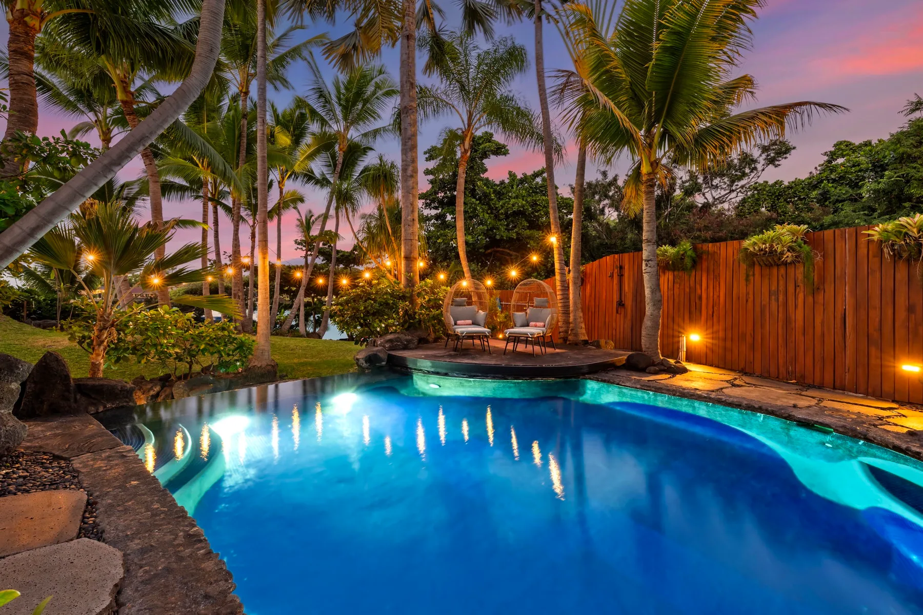 A swimming pool glows at dusk, surrounded by palm trees and outdoor seating.