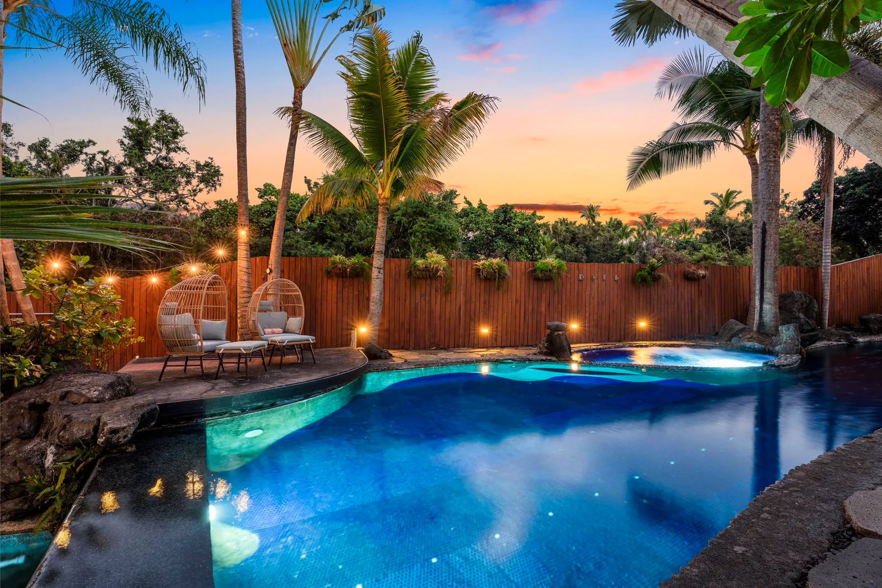 A swimming pool is surrounded by palm trees, seating, and a wooden fence at sunset.