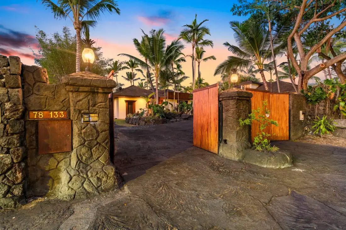 A tropical property entrance with stone gateposts and open wooden gates.