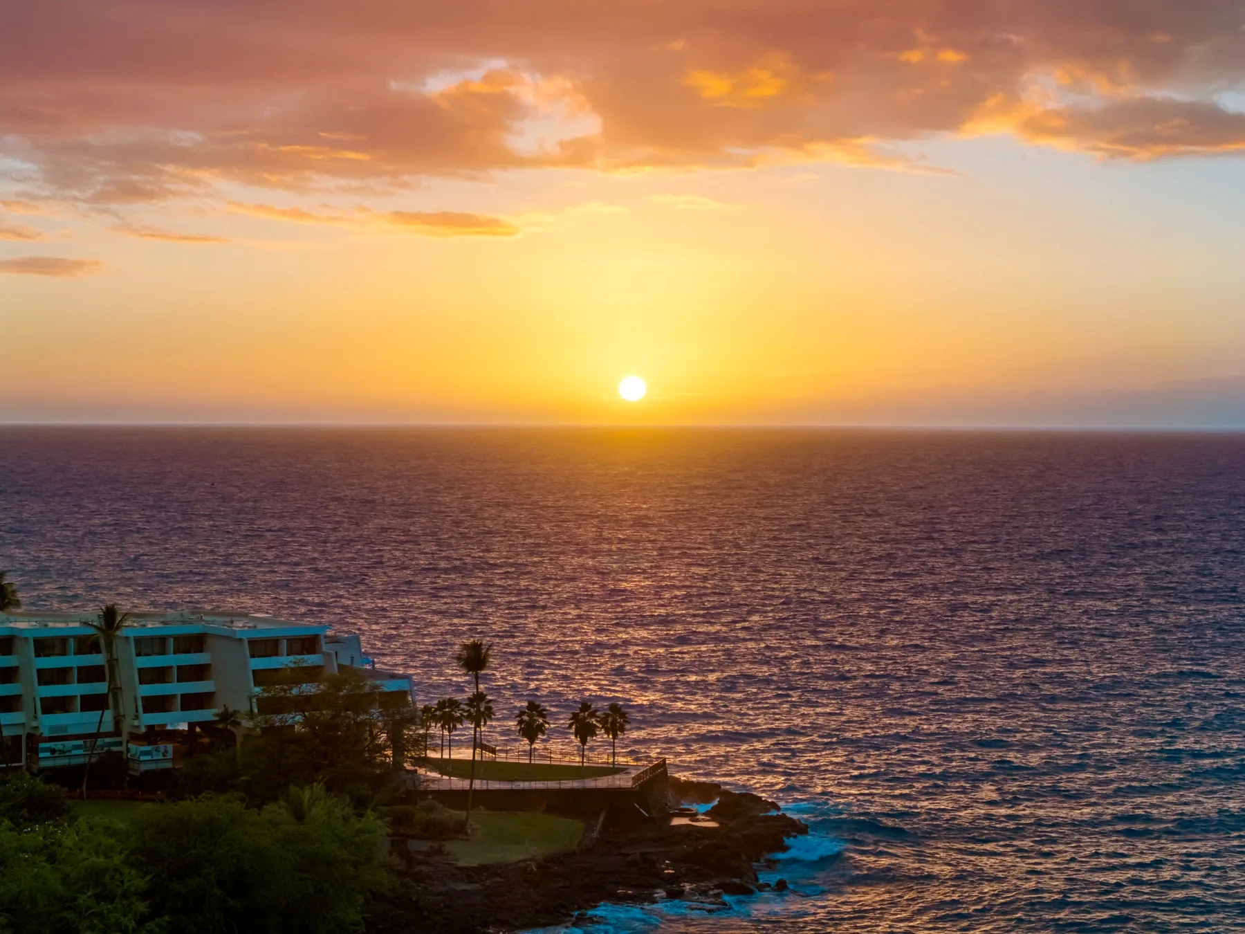 Sunset over the ocean, with a coastal building and palm trees in the foreground.