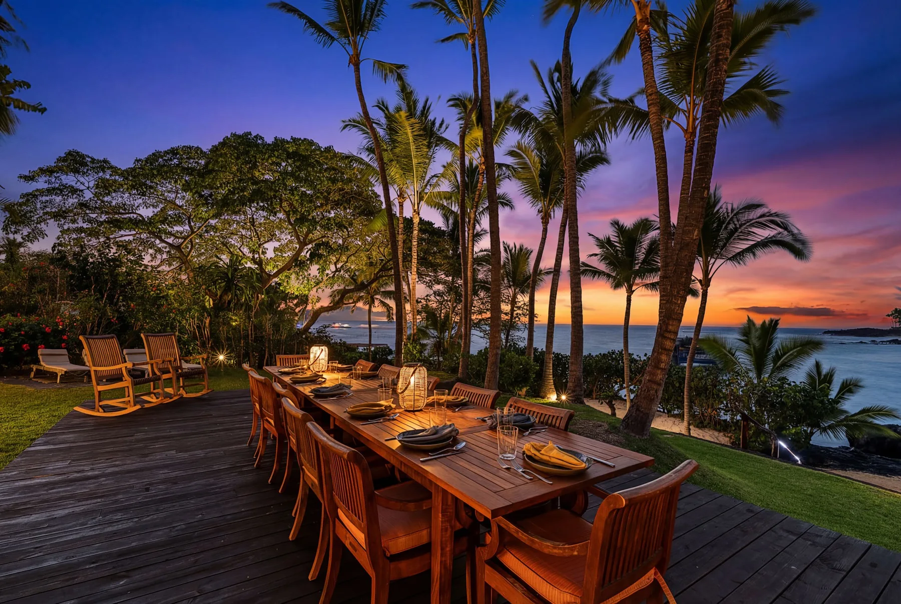 An outdoor dining area with a long wooden table overlooks the ocean at sunset.