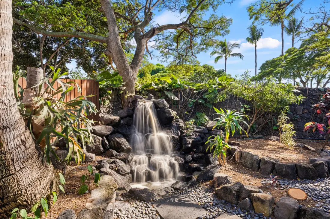 A garden waterfall flows over rocks beneath leafy trees.