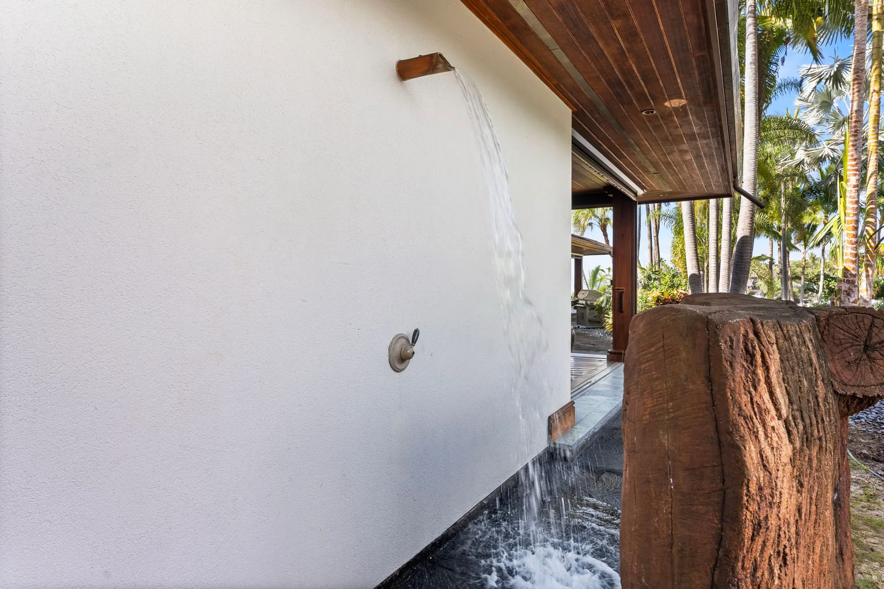 An outdoor shower runs beside a white wall under a wooden overhang.