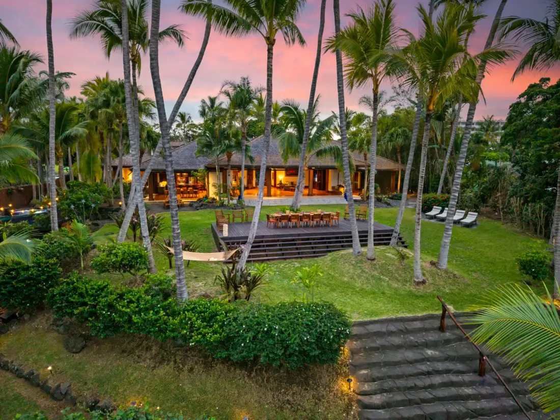 A tropical garden surrounds a warmly lit pavilion beside a lawn and stone steps.