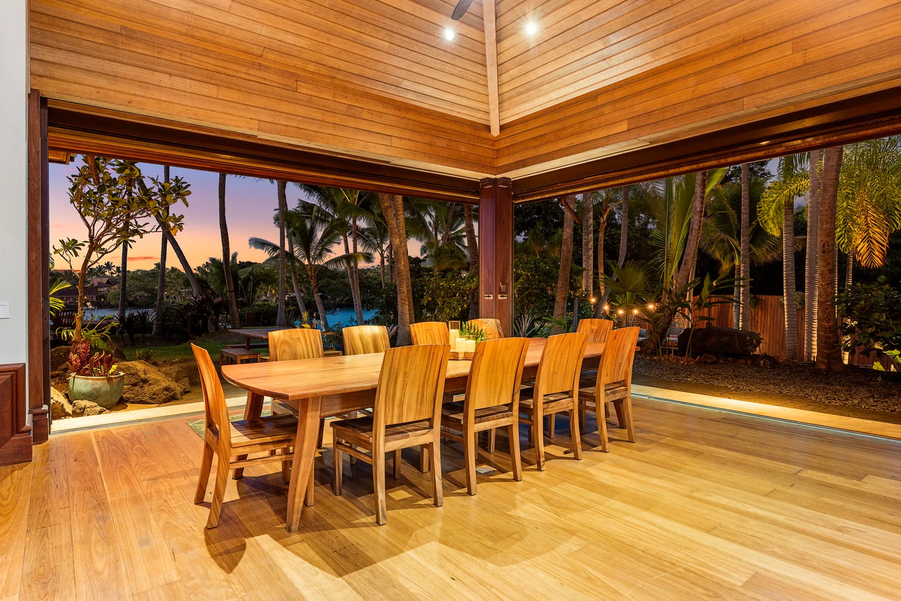 A dining area with a wooden table and chairs beside open views of tropical greenery.