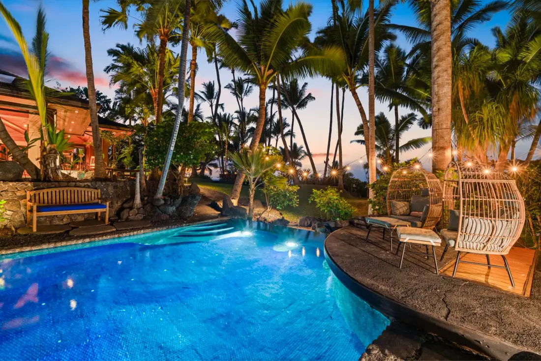 A swimming pool sits beside a curved deck, outdoor seating, and palm trees.