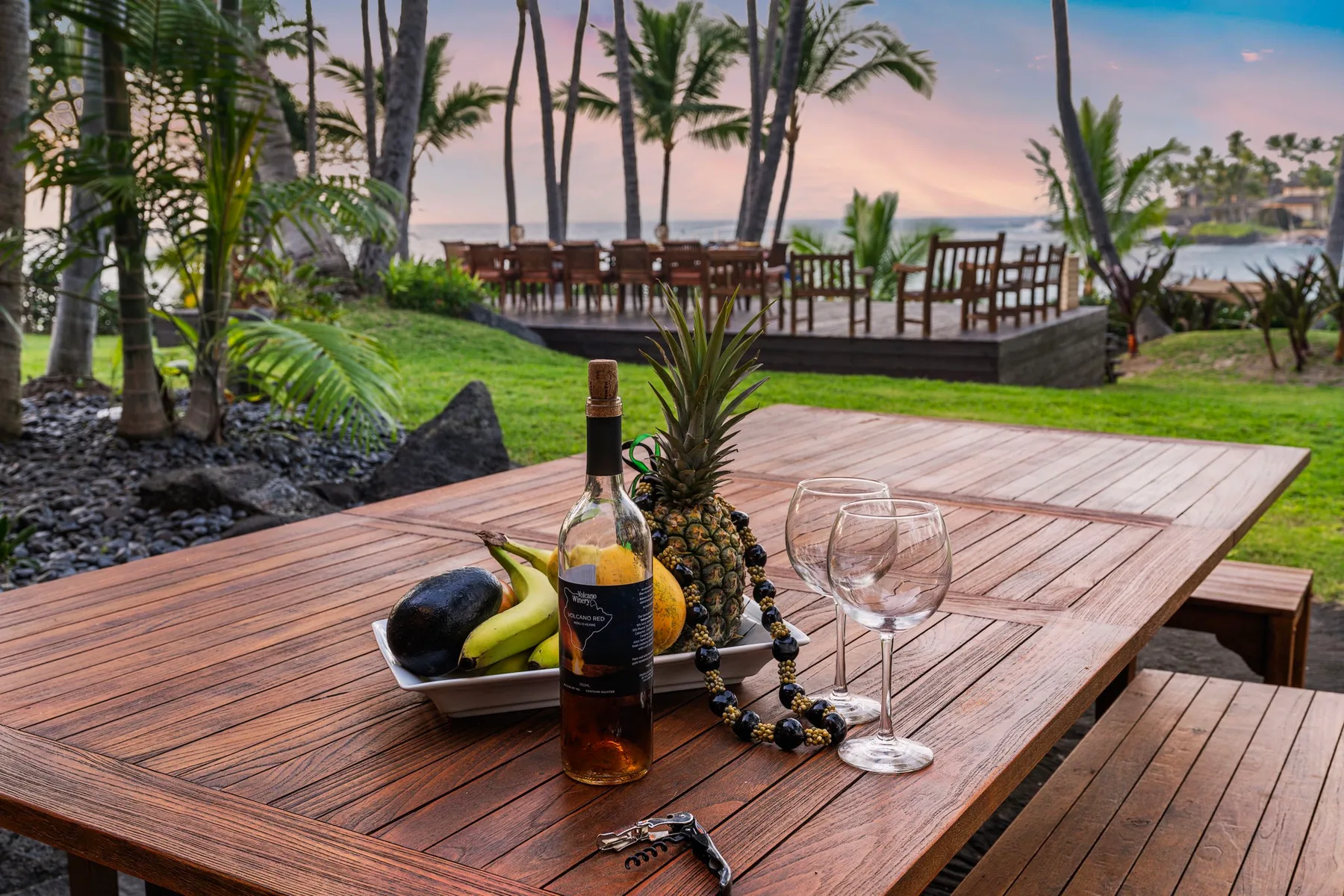 A wooden outdoor table with fruit and wine sits beside a tropical garden overlooking the water.