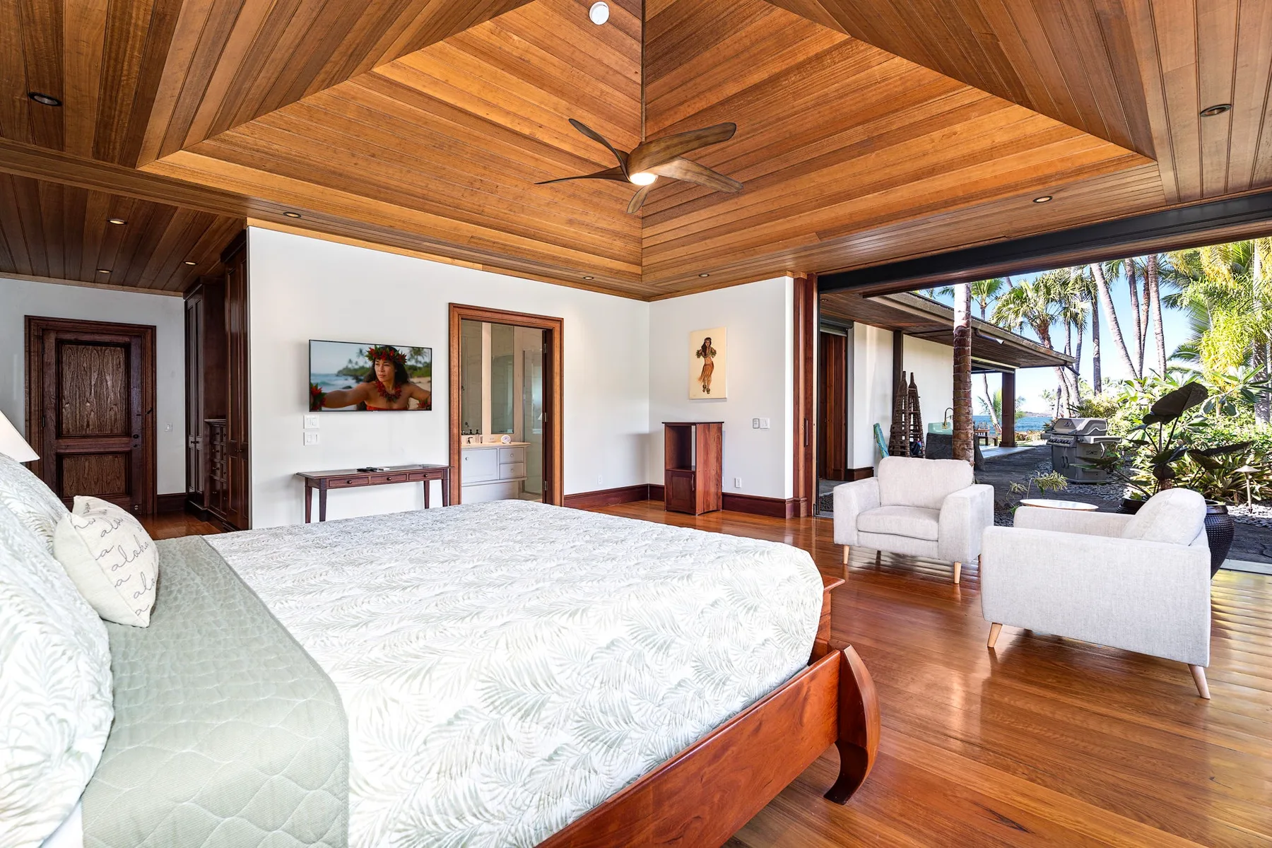 A bedroom with a large bed and an open sitting area overlooking palm trees.