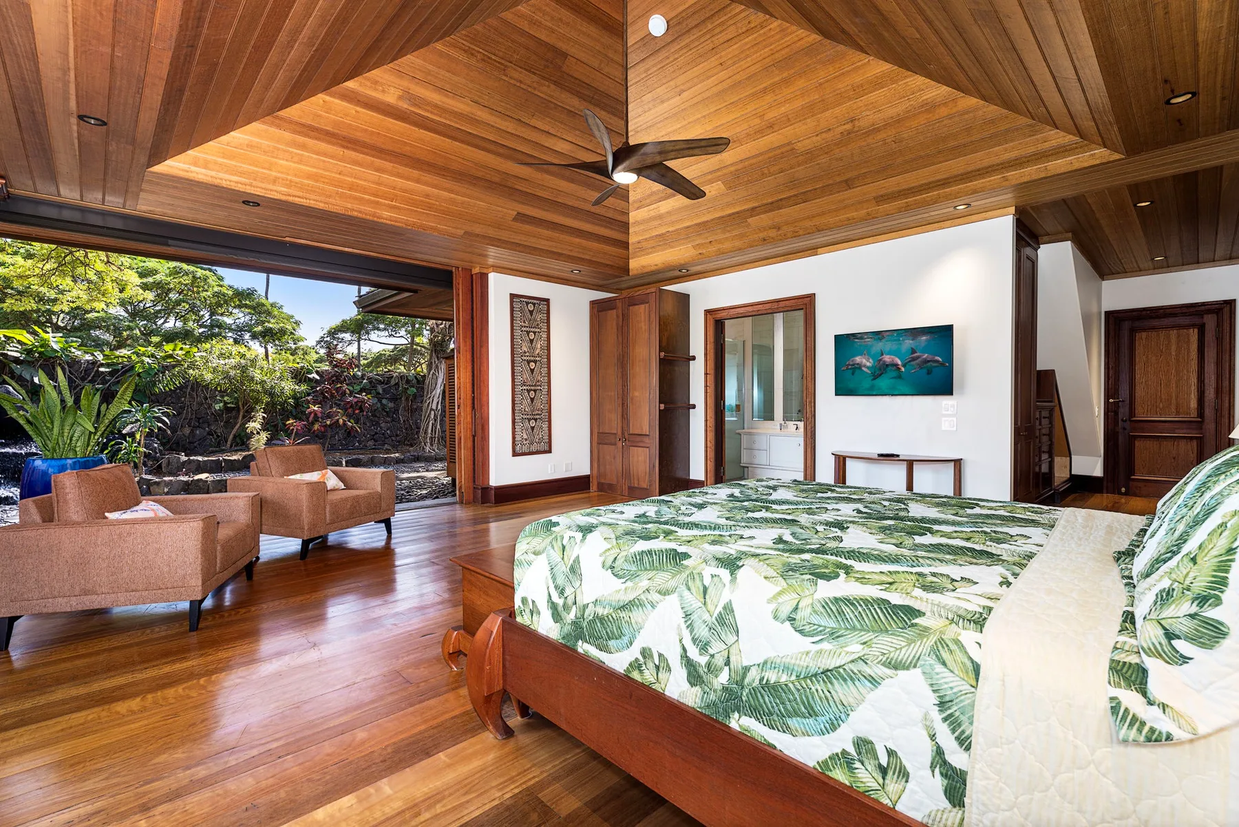 A bedroom with a leafy bedspread, wooden ceiling, and seating beside an open garden view.