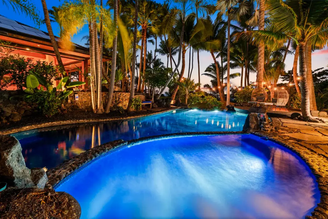 A glowing swimming pool sits among palm trees and tropical landscaping at night.