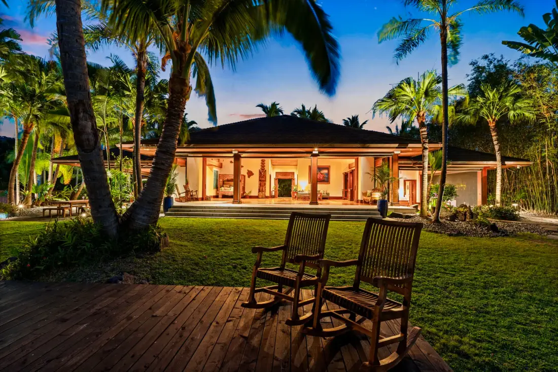 A tropical house faces a lawn with palm trees and wooden seating.