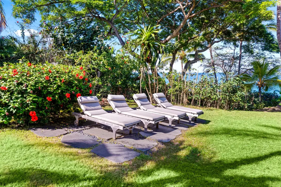 A garden seating area with lounge chairs sits beside flowering shrubs and trees.