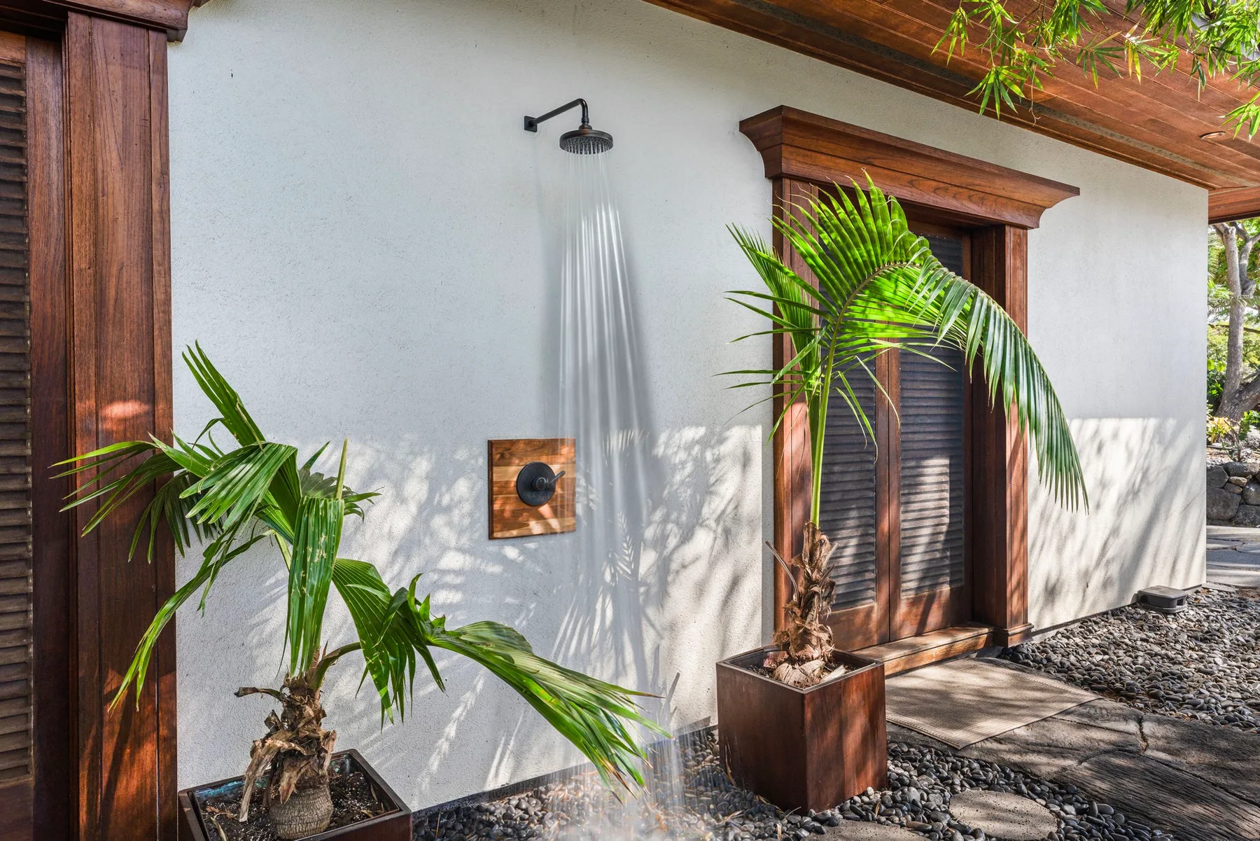 An outdoor shower is mounted on a white wall beside wooden shutters and potted palms.