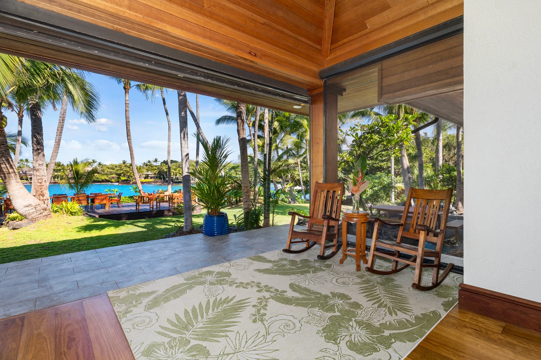 A covered patio with wooden seating overlooks a palm-lined garden and pool.