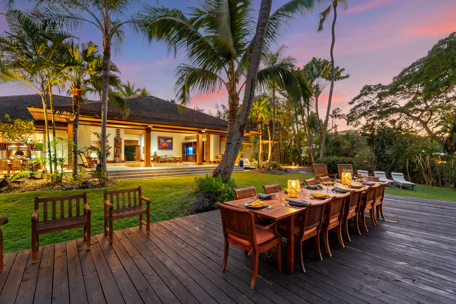 An outdoor dining area on a wooden deck faces a warmly lit building amid tropical trees.