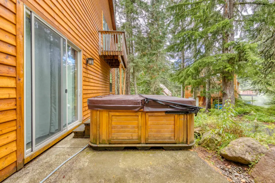 An outdoor hot tub sits beside a wood-sided house amid trees.