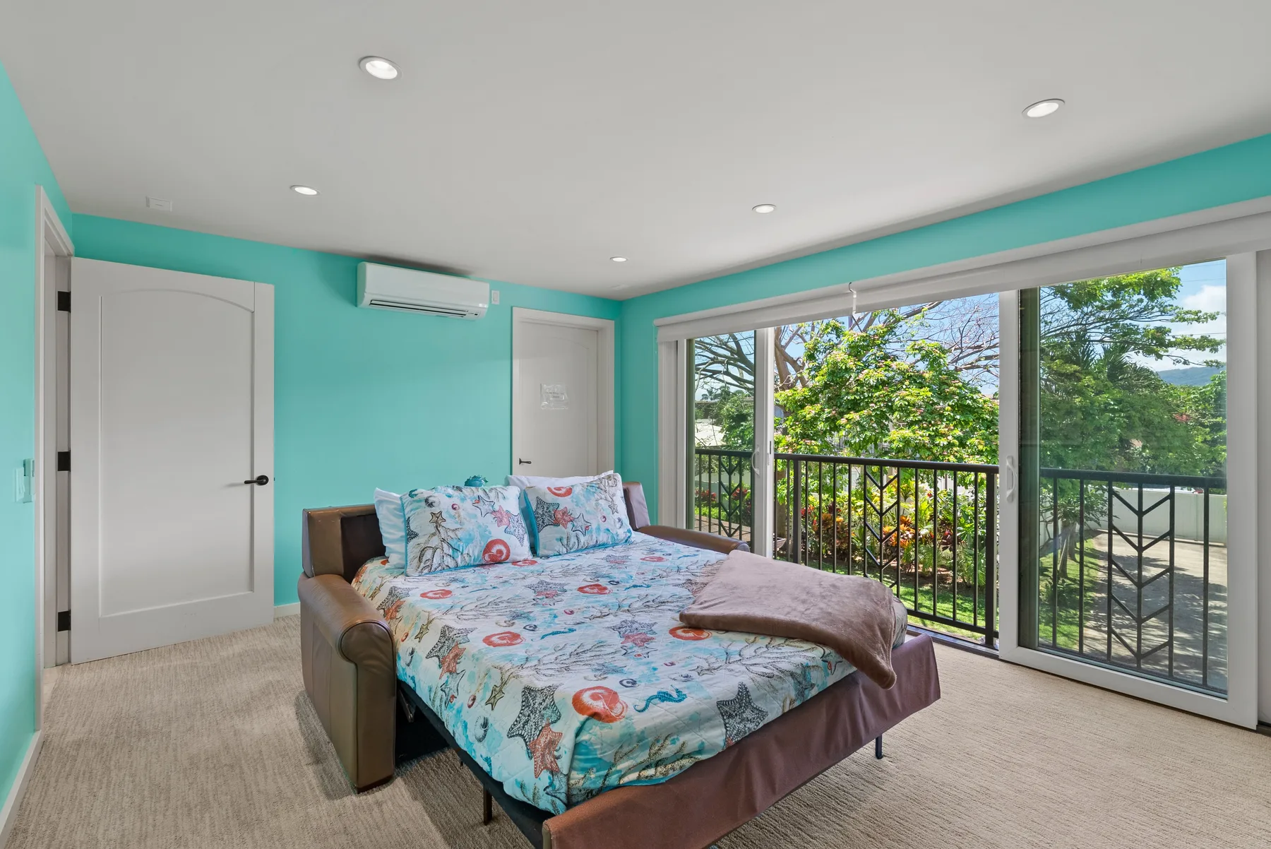 Un dormitorio con una cama junto a grandes ventanas con vista a árboles y una baranda de balcón.