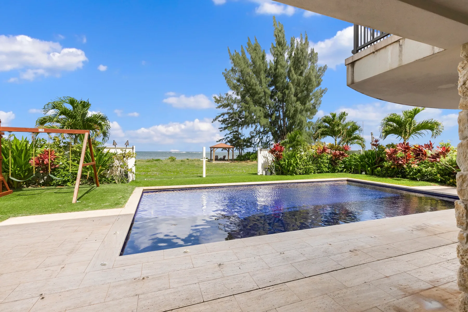 Una piscina y un patio pavimentado dan a un jardín ajardinado y al mar.