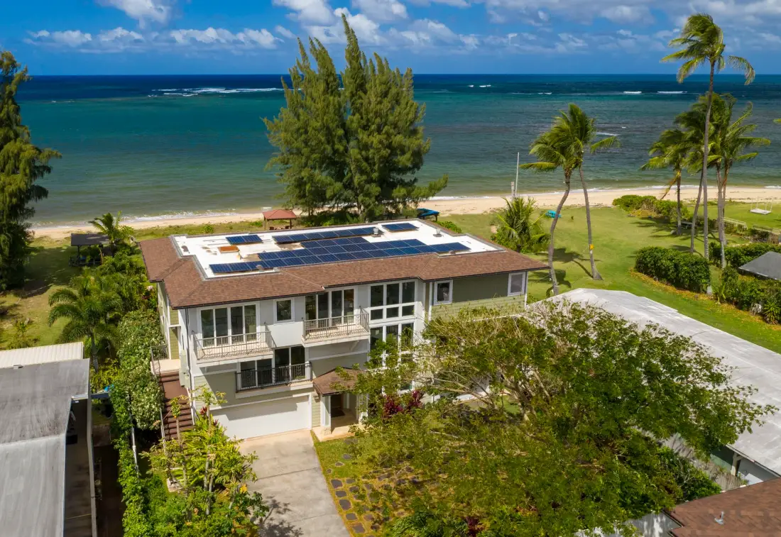 Una casa costera con paneles solares está junto a una playa de arena y aguas turquesas.