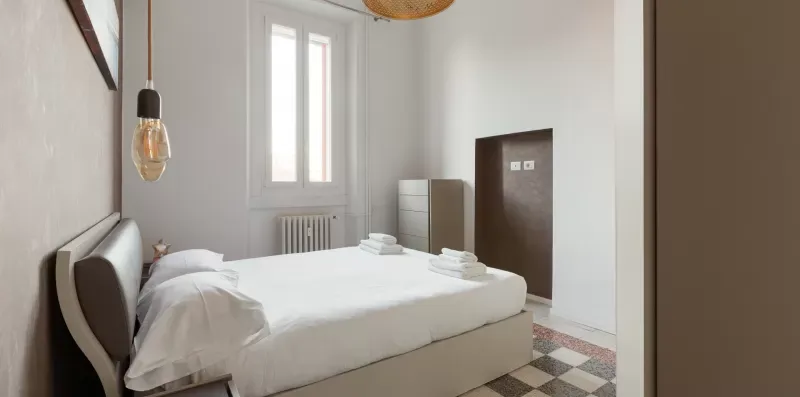 A bedroom with a neatly made bed, tall windows, and a small dresser.