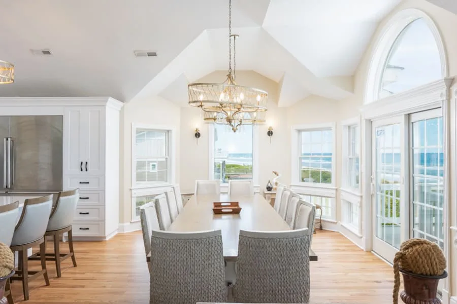 A bright dining room has a long table beside windows overlooking the water.