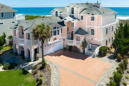 A large pink coastal house stands beside a paved driveway, with the ocean visible behind it.