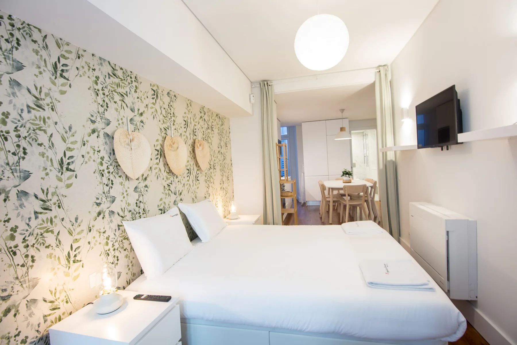 A bright bedroom with a bed and a dining area visible beyond the curtains.