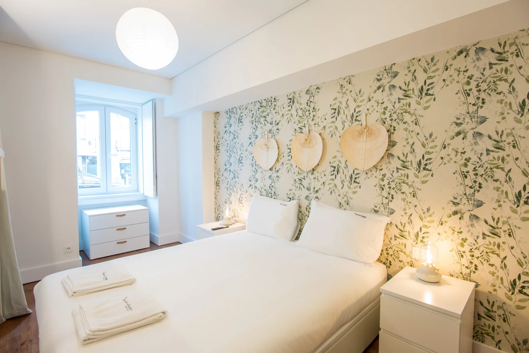 A bright bedroom with a white bed and leafy patterned wallpaper.