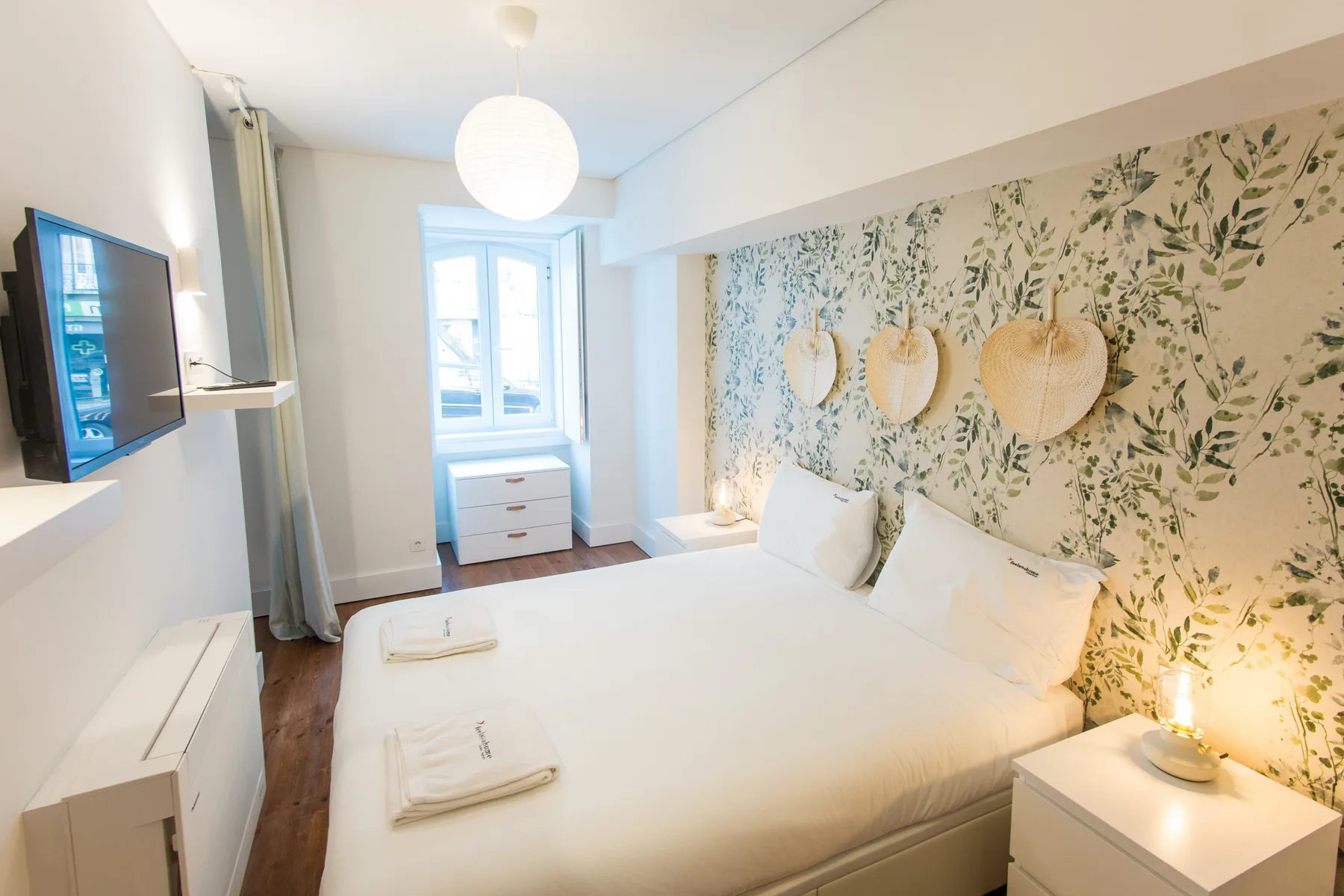 A bright bedroom with a large bed and leafy patterned wallpaper.