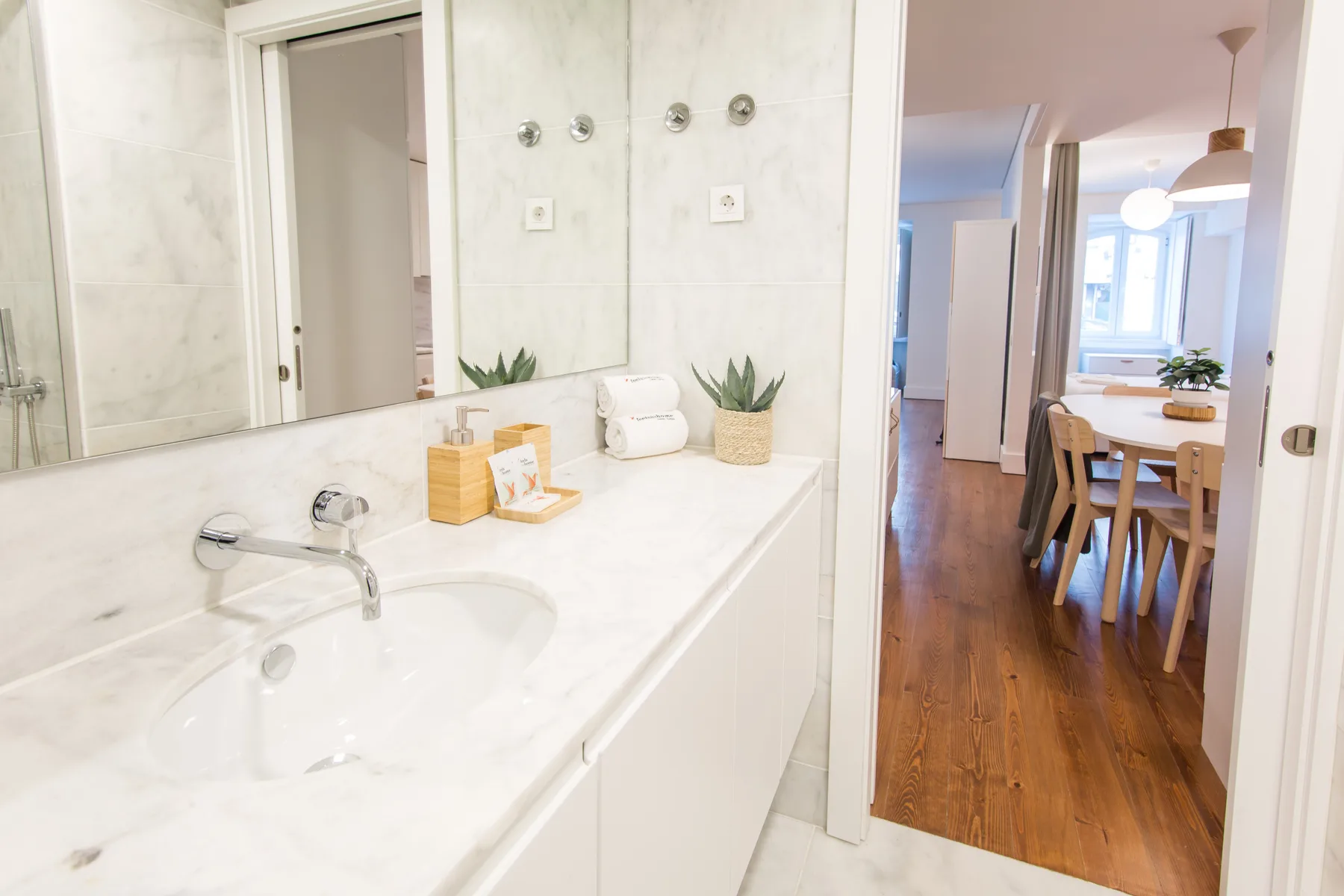 A bright bathroom vanity opens toward a dining area with a wooden table.