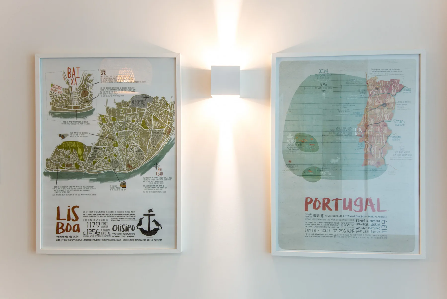 Two framed maps hang on a wall beside a lit wall sconce.