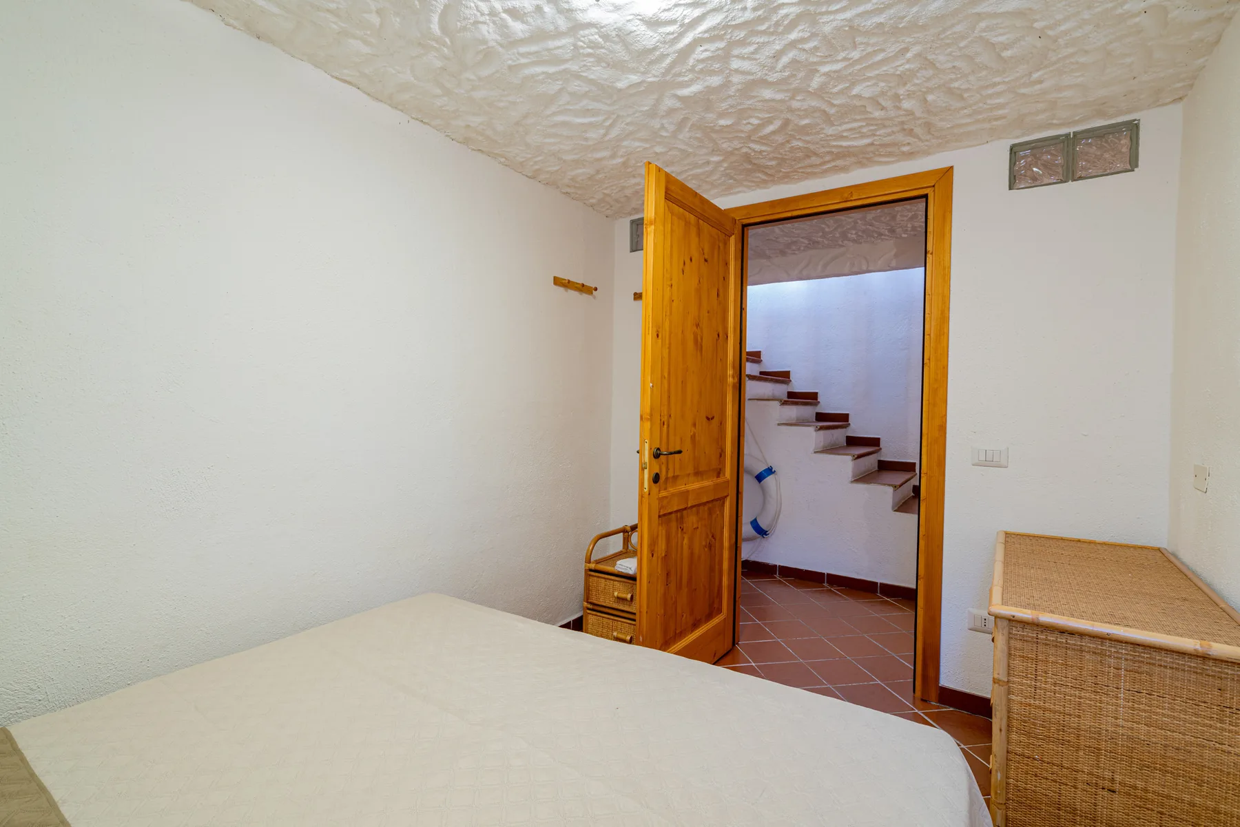 Un dormitorio con una cama, un baúl de mimbre y una puerta abierta hacia unas escaleras.