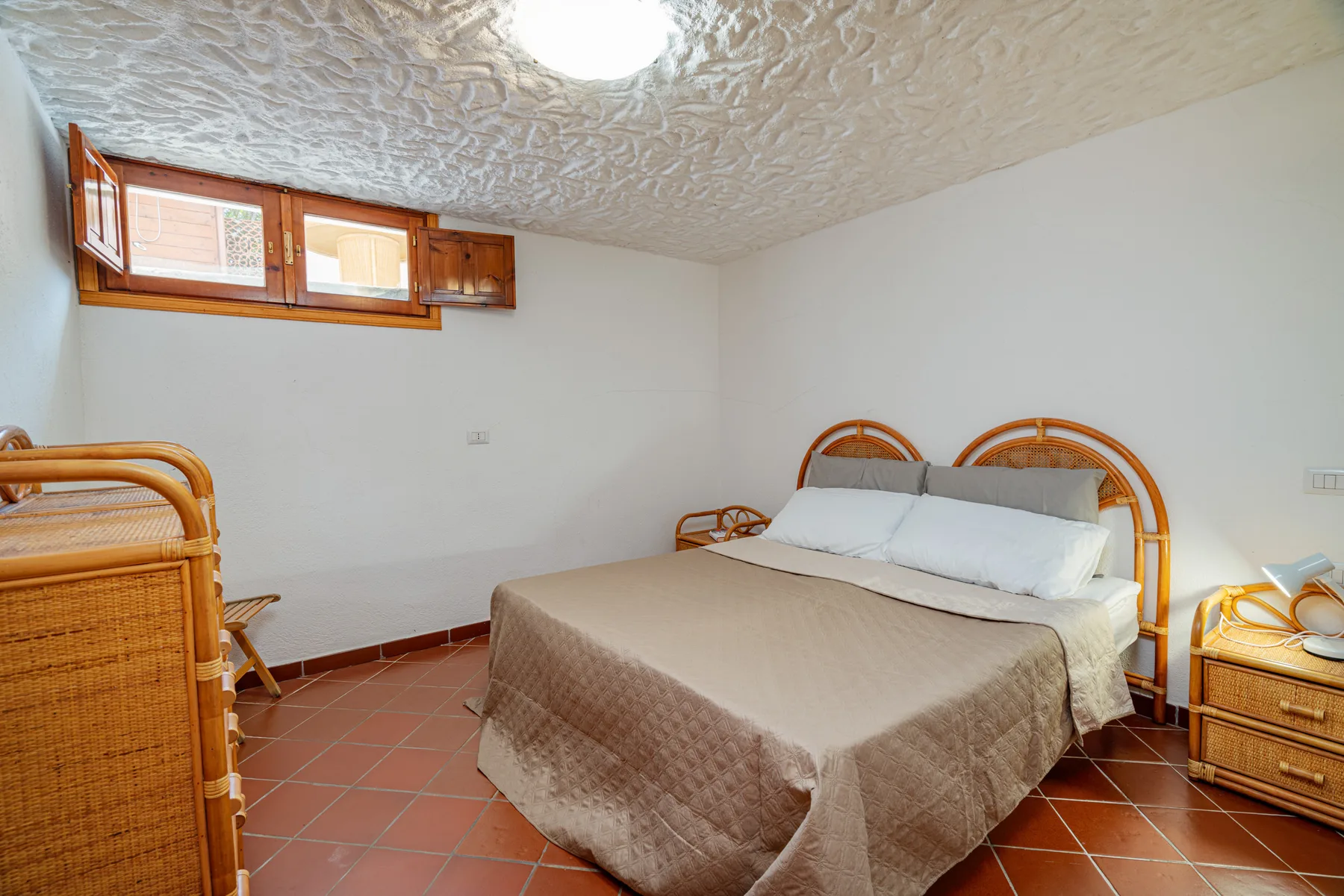 Un dormitorio con una cama, asientos de mimbre y ventanas altas.