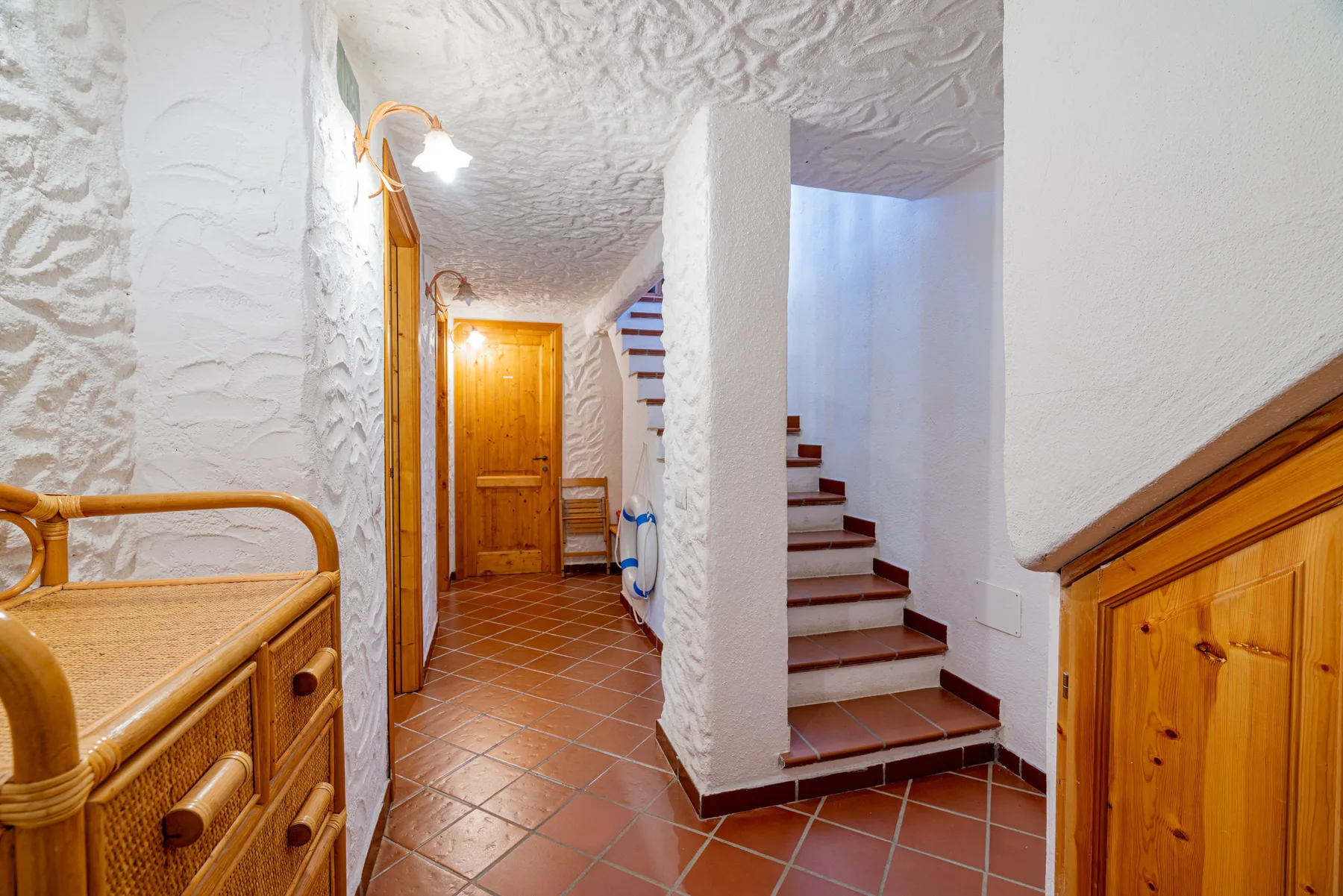 Un pasillo interior de paredes blancas, suelo de baldosas color terracota y una escalera.