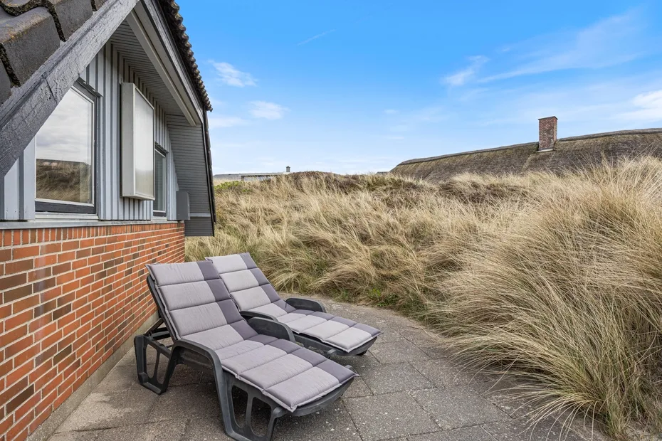 An outdoor patio with reclining seating beside a house and grassy dunes.
