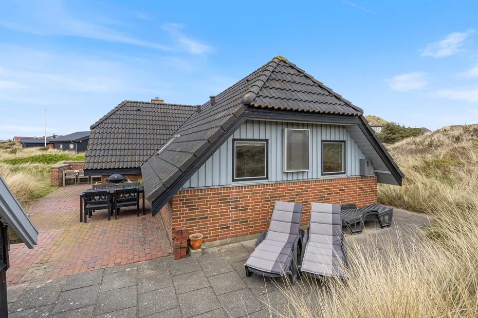 A brick house with a sloped roof and outdoor seating beside sandy dunes.
