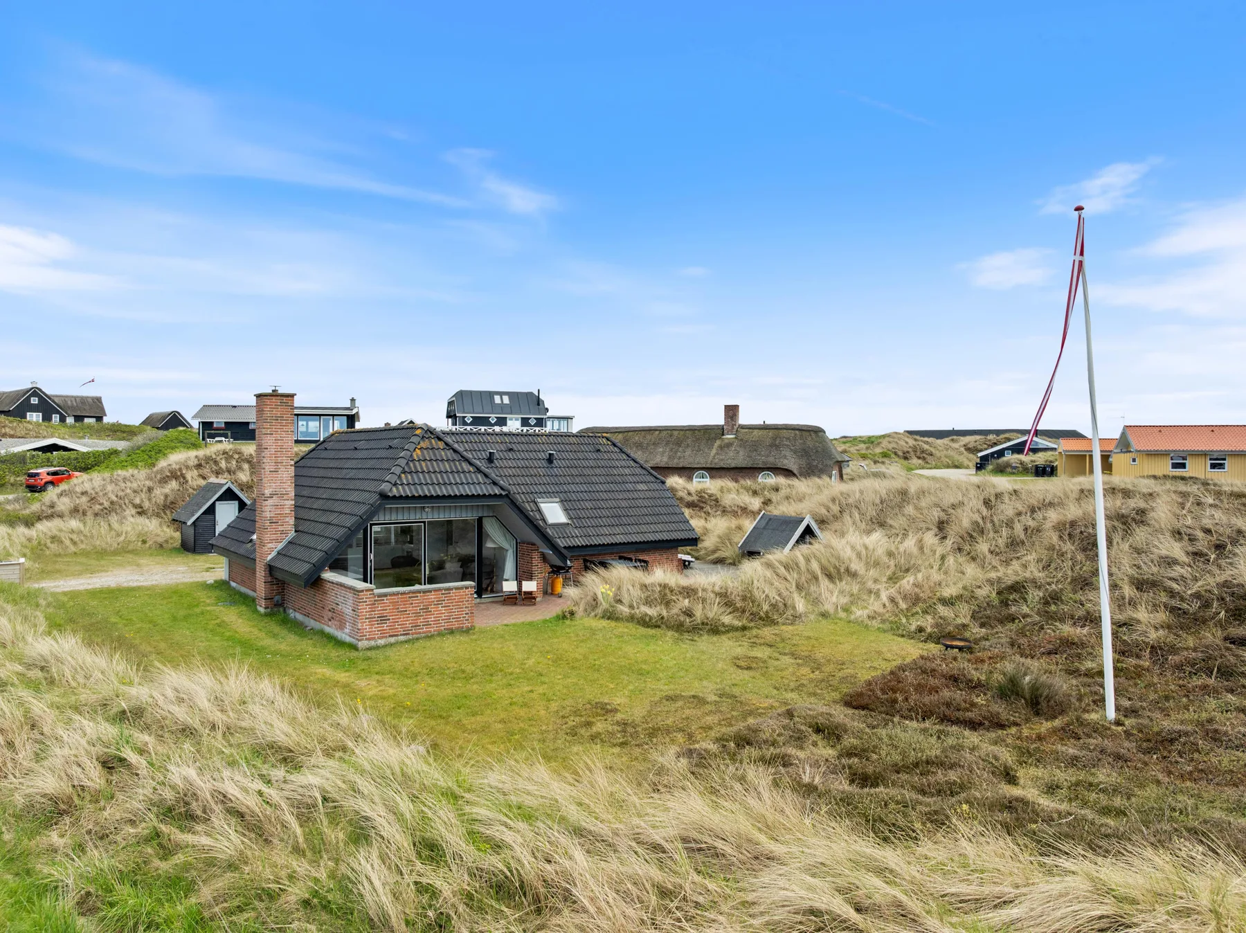 A low brick house with a dark roof sits among grassy dunes beneath a blue sky.