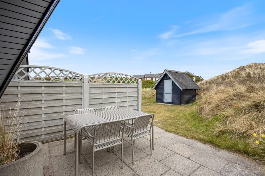A paved patio with a dining table sits beside a fenced yard and grassy dune.