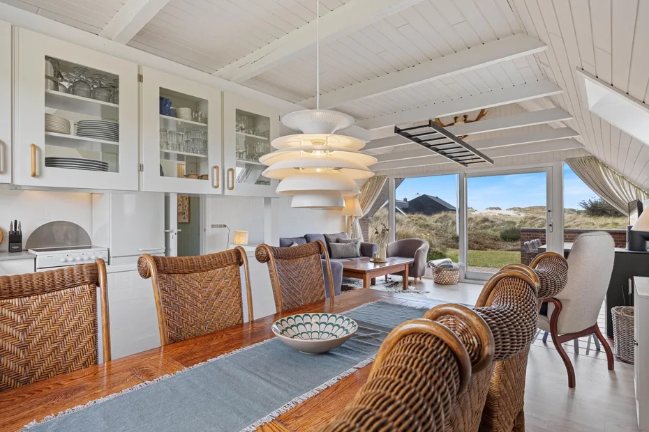 A bright dining area has woven seating, a long table, and wide windows overlooking the landscape.