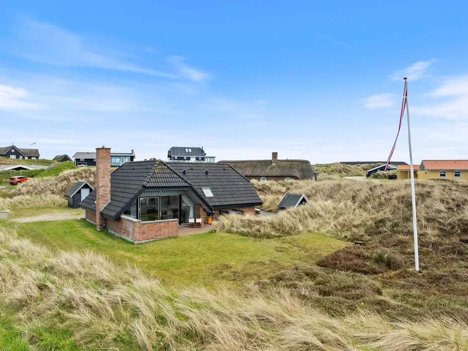 A low-roofed house sits among grassy dunes beneath a clear blue sky.