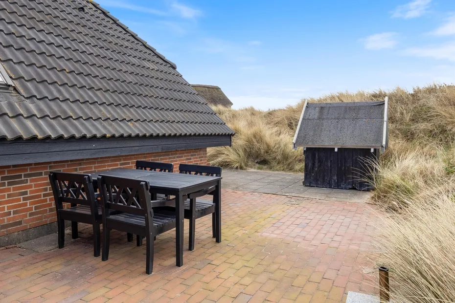 A paved outdoor patio with dark seating beside a brick building and grassy dunes.