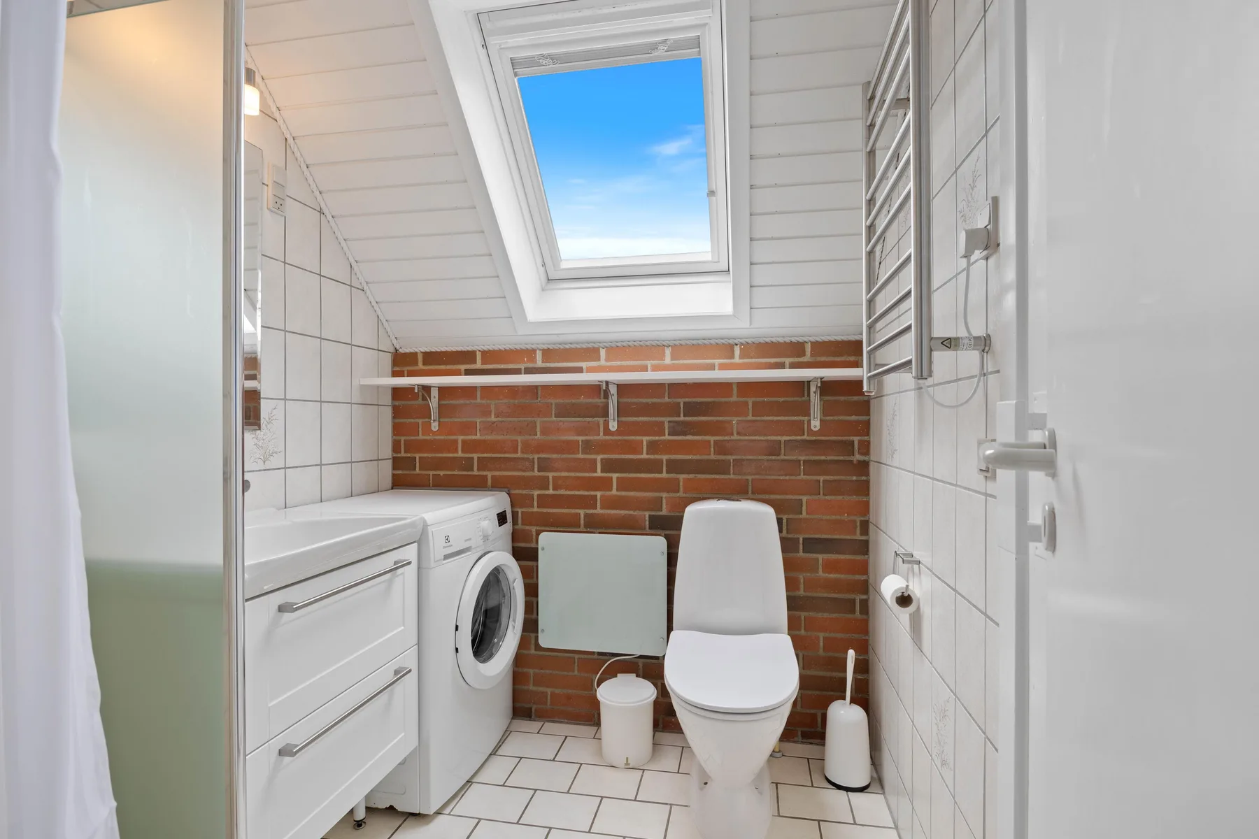 A tiled bathroom with a toilet and washing machine beneath a skylight.