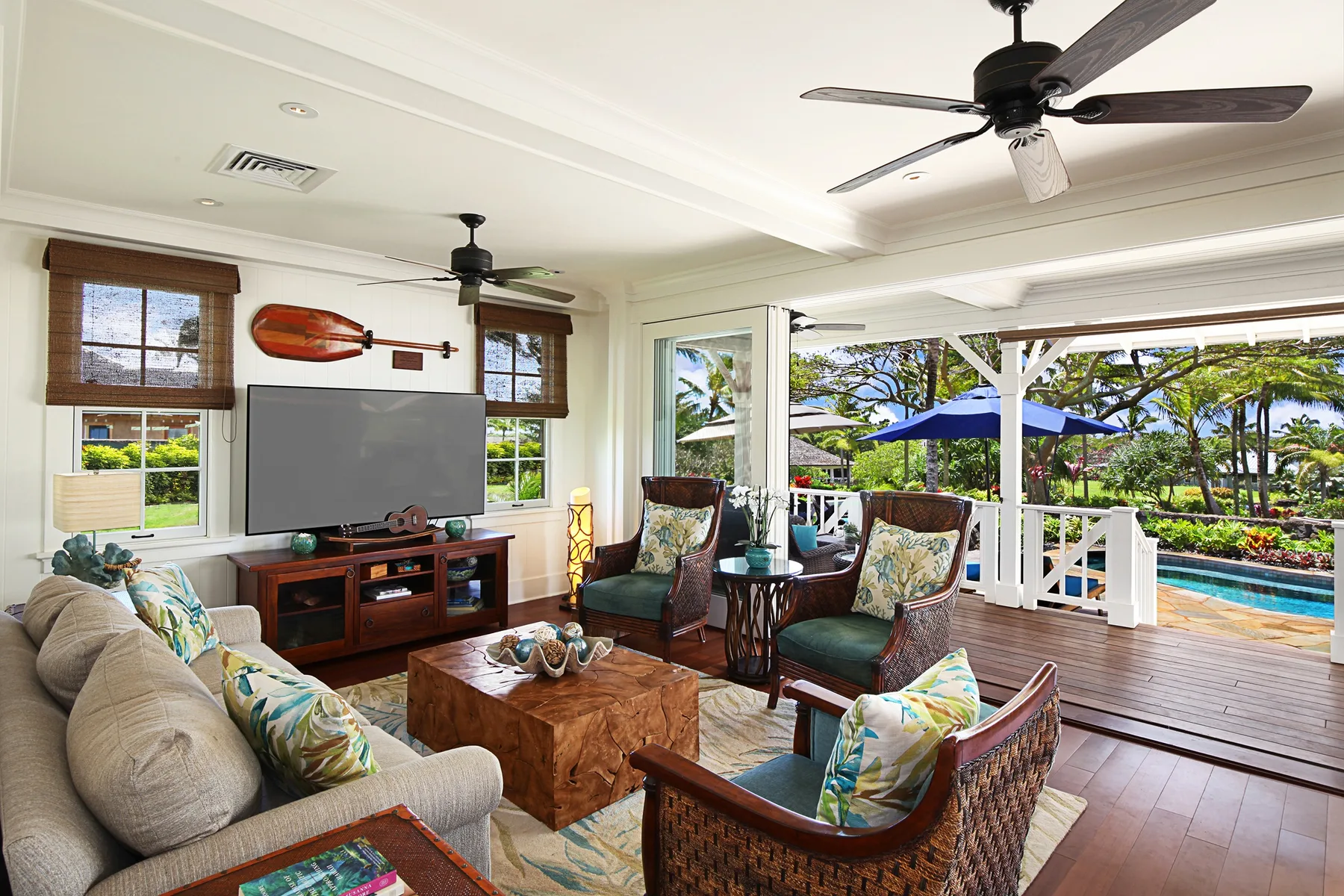 A bright living room opens onto a covered deck with garden and pool views.
