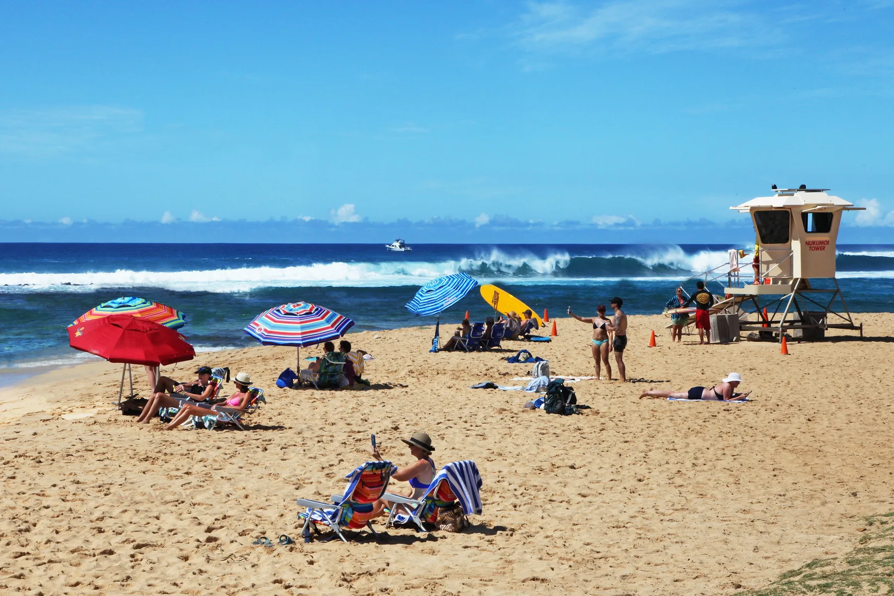 Beachgoers relax on a sandy beach beneath colorful umbrellas, with waves in the background.