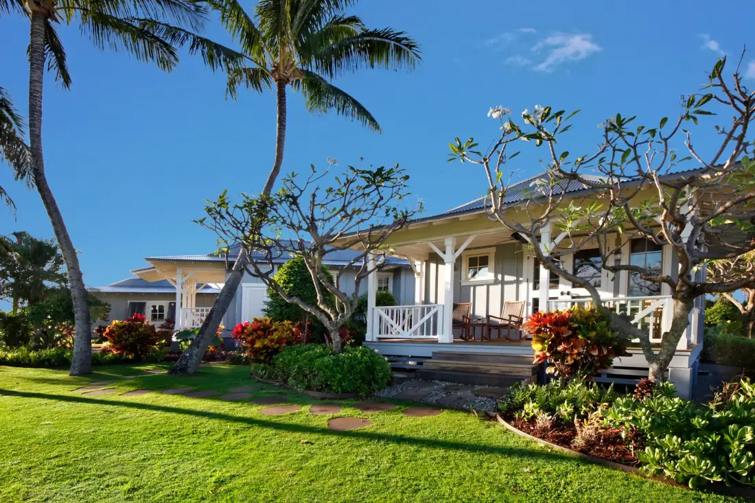 A white house with a covered porch stands behind a green lawn and tropical plants.