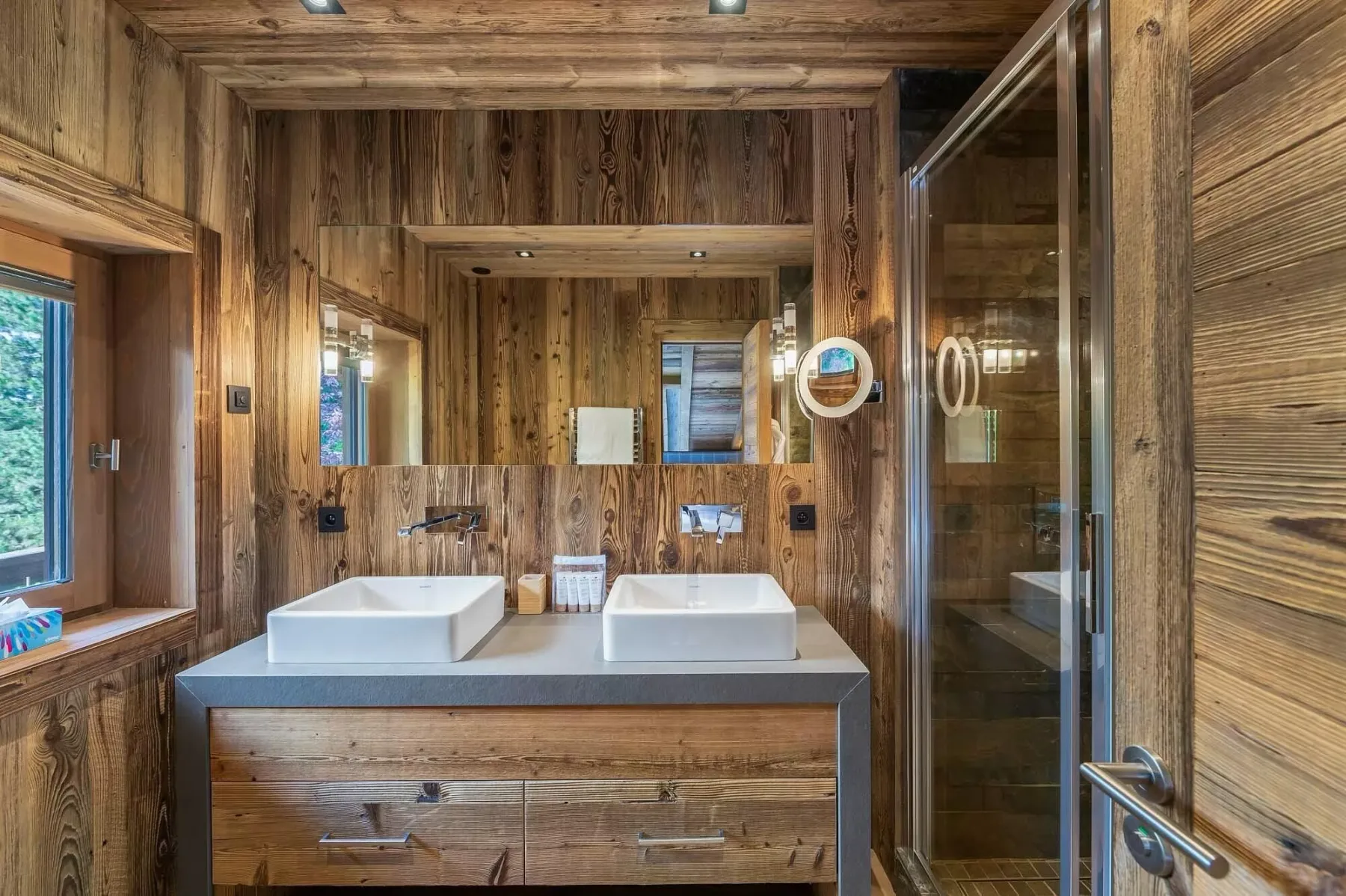 Baño revestido de madera con dos lavabos y una ducha de vidrio.